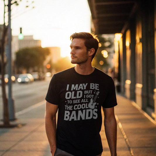 Male model leaning against a brick wall wearing a black crew-neck t-shirt with a vintage guitar graphic and “I May Be Old But I Got To See All The Cool Bands” text, lifestyle street shot