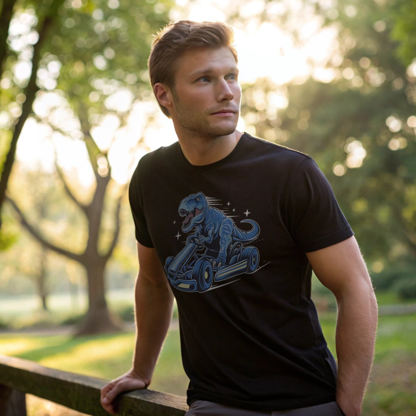 Male model leaning on a wooden fence in a black crew-neck t-shirt with a blue dinosaur kart graphic, lifestyle outdoor shot.