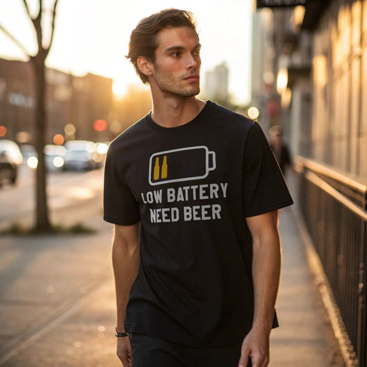 Male model walking an urban sidewalk in a black crew-neck t-shirt with a low battery icon and “Low Battery Need Beer” text, lifestyle street shot.