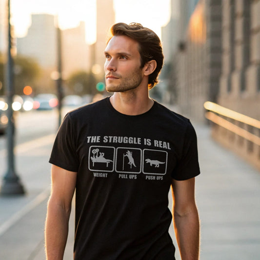 Male model strolling an urban sidewalk in a black crew-neck t-shirt with “The Struggle Is Real” graphic, lifestyle street shot.