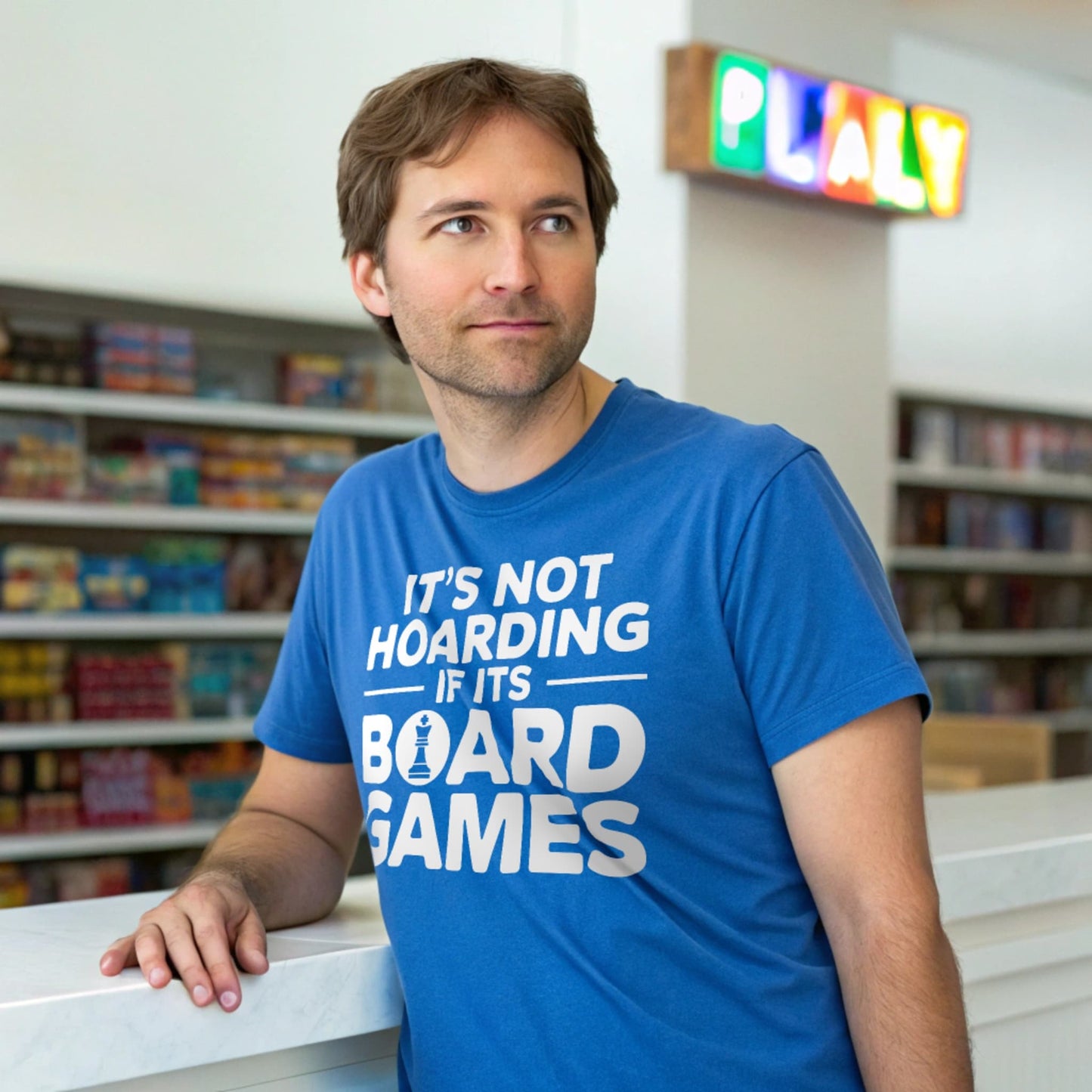 Hyper-realistic mockup of a 25–40-year-old man wearing a royal blue Board Games Hoarder T-Shirt leaning against a board-game boutique counter with neon ‘PLAY’ sign and shelves behind him.