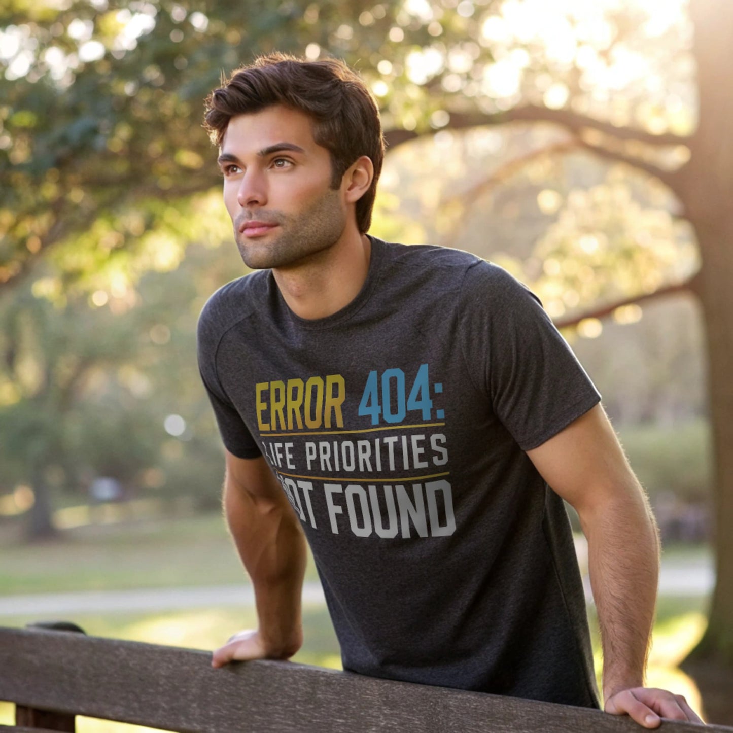 Male model leaning on a park bench in a dark heather crew-neck t-shirt with “Priorities Not Found” tech graphic, outdoor lifestyle shot in morning light.