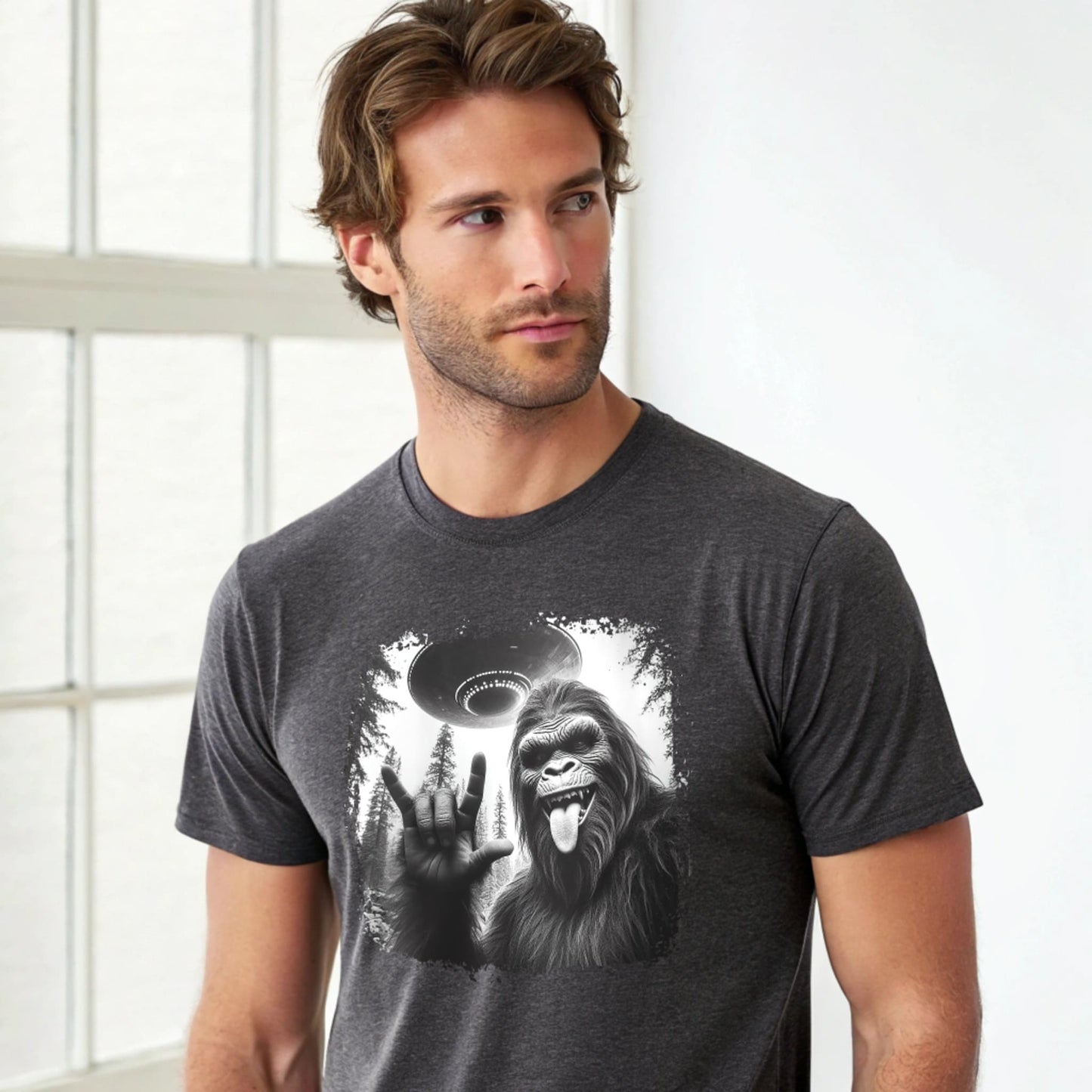 Male model wearing a dark heather crew-neck t-shirt featuring a white silhouette of Sasquatch raising its hand graphic, studio shot by a window-lit backdrop.