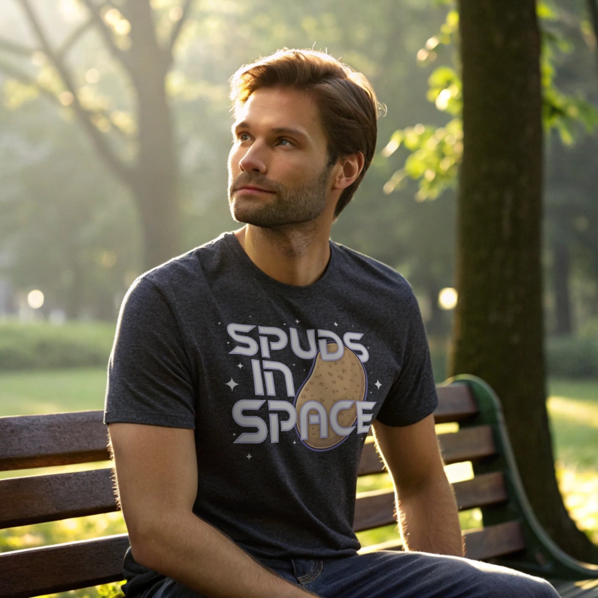Male model seated on a park bench in a dark heather crew-neck t-shirt with “Spuds in Space” graphic, outdoor lifestyle shot.