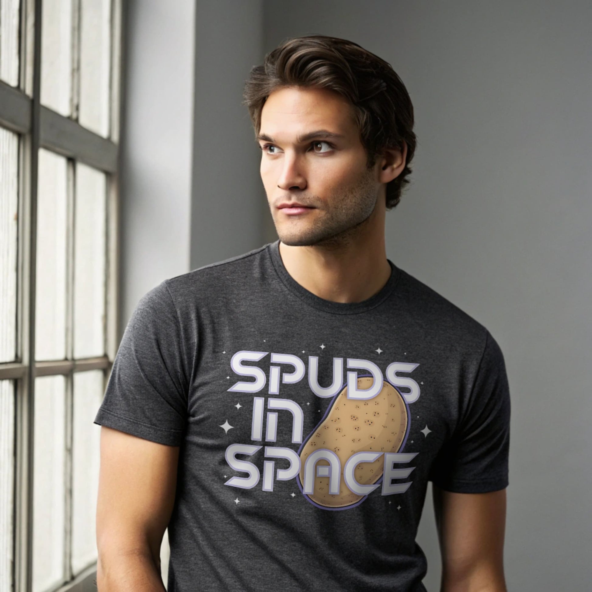 Male model wearing a dark heather crew-neck t-shirt with “Spuds in Space” graphic, studio shot by a window-lit backdrop.