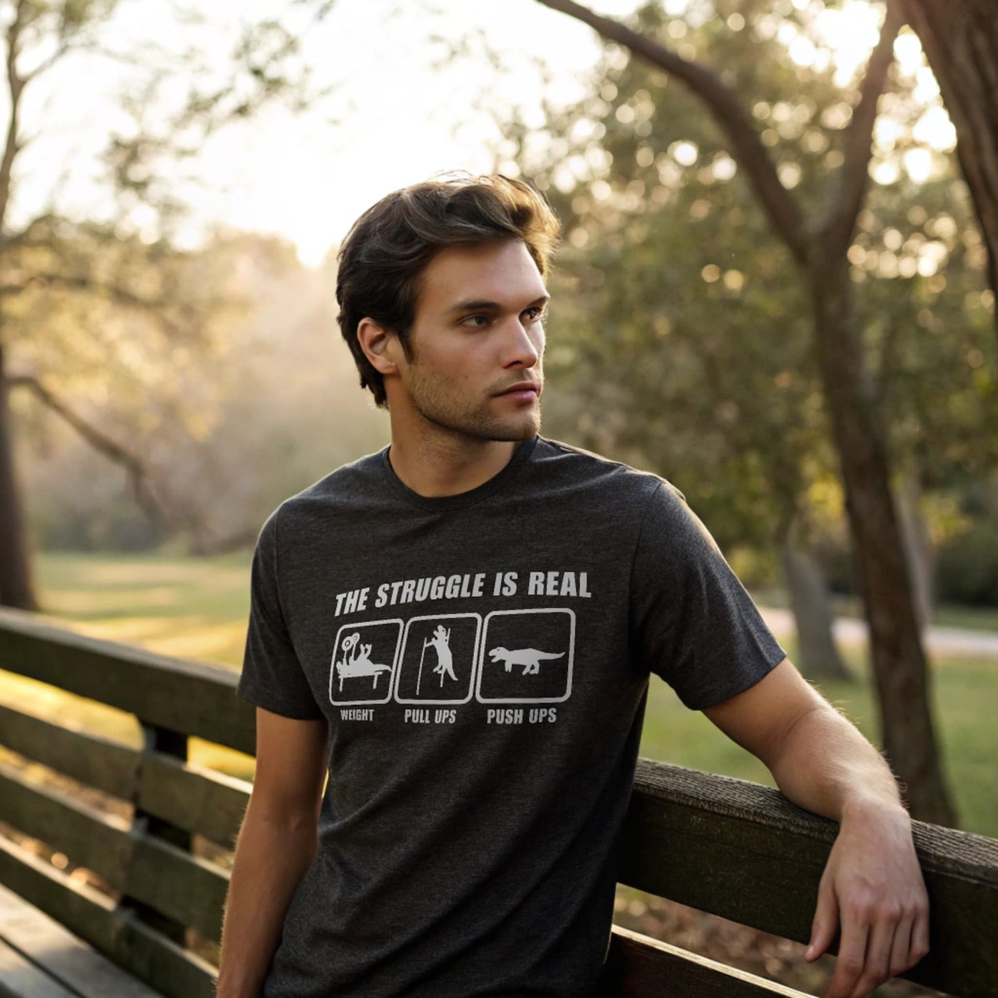 Male model leaning on a park bench in a dark heather crew-neck t-shirt with “The Struggle Is Real” graphic, lifestyle outdoor shot.
