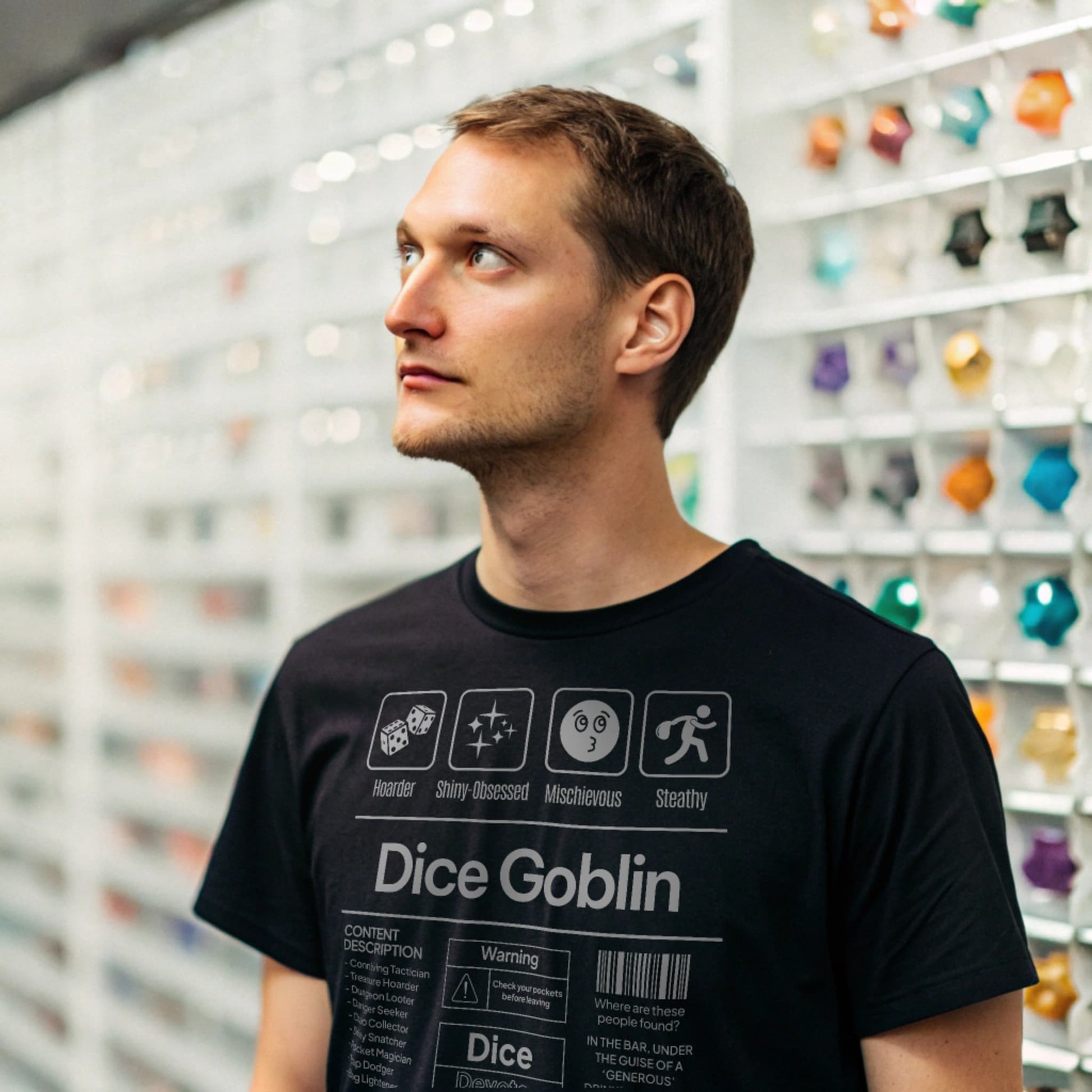 Black lifestyle mockup of Dice Goblin T-Shirt on a male model browsing a glass dice display in a specialty dice shop