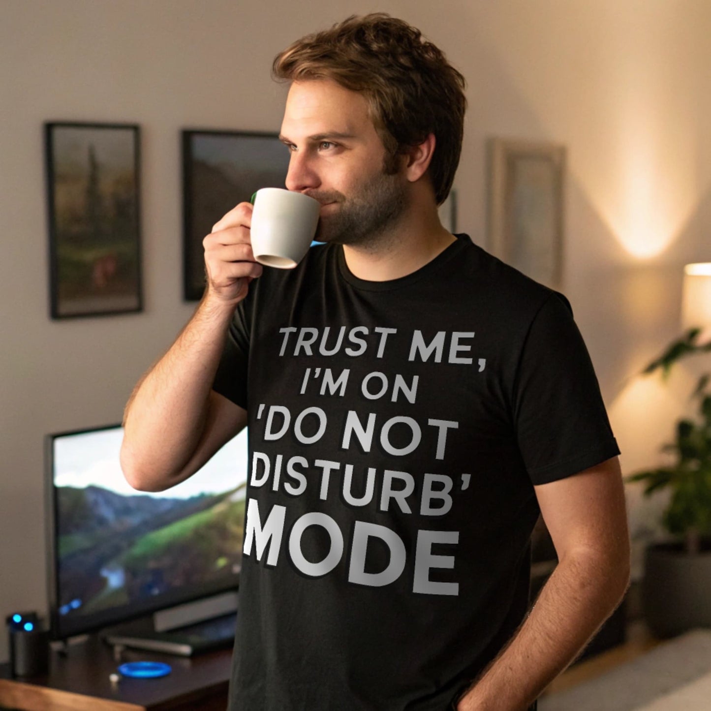 Realistic mockup of man drinking coffee while wearing a black “Do Not Disturb Mode” shirt in cozy gaming room