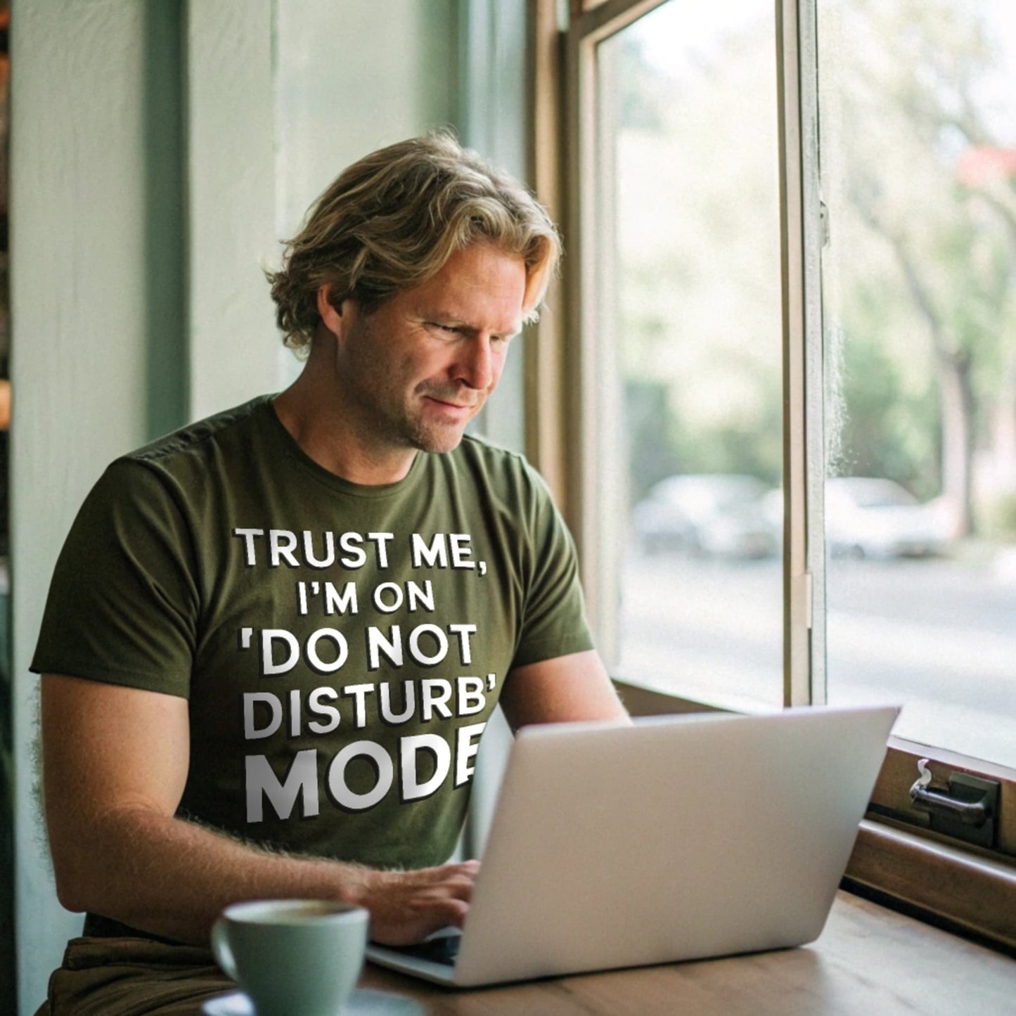 Realistic mockup of man working on laptop in a cafe while wearing a military green “Do Not Disturb Mode” shirt