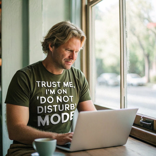 Realistic mockup of man working on laptop in a cafe while wearing a military green “Do Not Disturb Mode” shirt