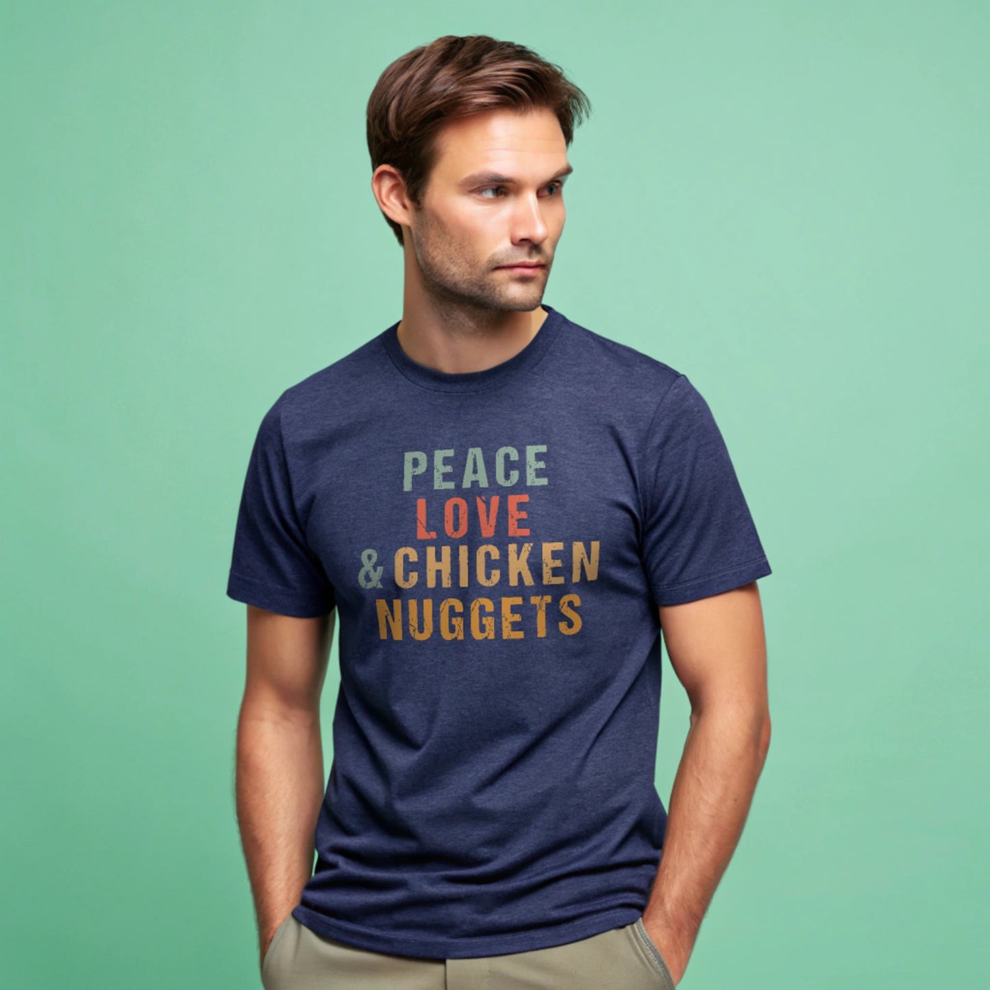 Male model wearing a heather navy crew-neck t-shirt with “Peace Love & Chicken Nuggets” text in multicolor block letters, studio shot on a mint-green background.