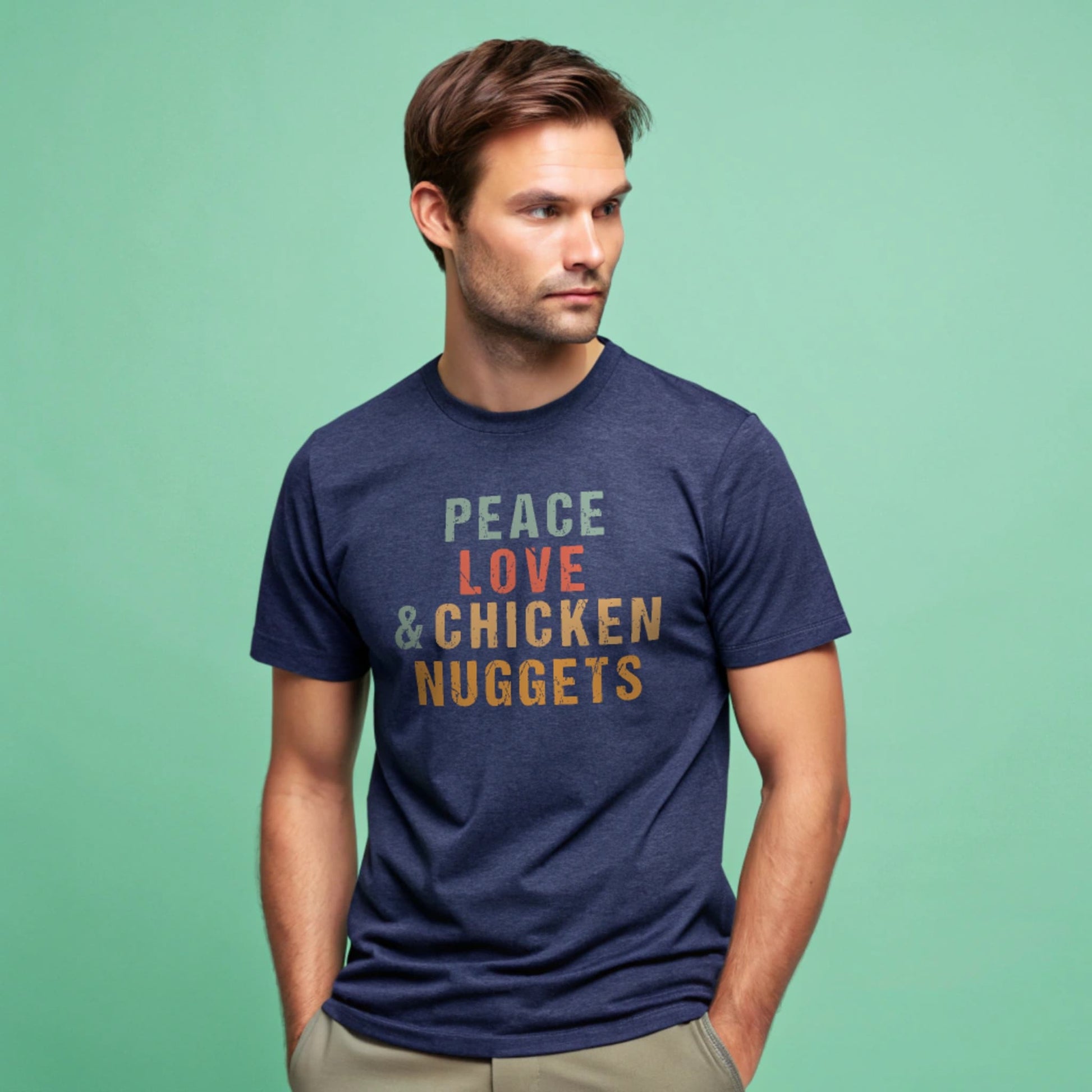 Male model wearing a heather navy crew-neck t-shirt with “Peace Love & Chicken Nuggets” text in multicolor block letters, studio shot on a mint-green background.