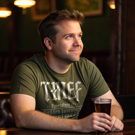 Man in his 30s sitting at a bar wearing a military green “Thief: Specializes in Sudden Inventory Adjustments” T-shirt, holding a beer and smirking off to the side