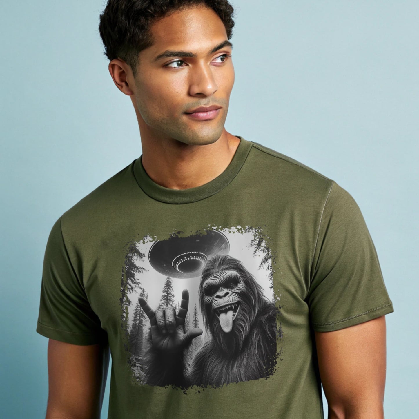 Male model wearing a military green crew-neck t-shirt featuring a white silhouette of Sasquatch raising its hand graphic, studio shot on a pale blue backdrop.