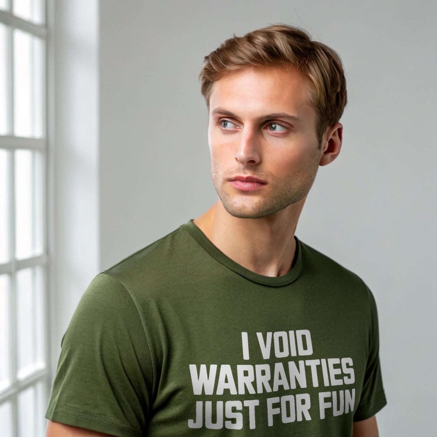 Male model wearing a plain military green Gildan 64000 crew-neck t-shirt with “I VOID WARRANTIES JUST FOR FUN” text, studio shot on light gray background.