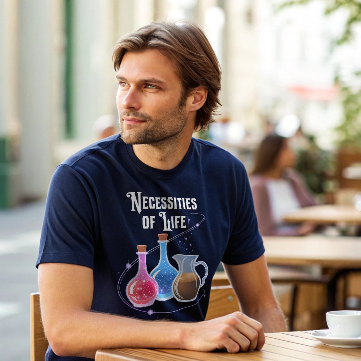 Male model seated at an outdoor cafĆ© table in a navy crew-neck t-shirt with āNecessities of Lifeā potion graphic, lifestyle shot.