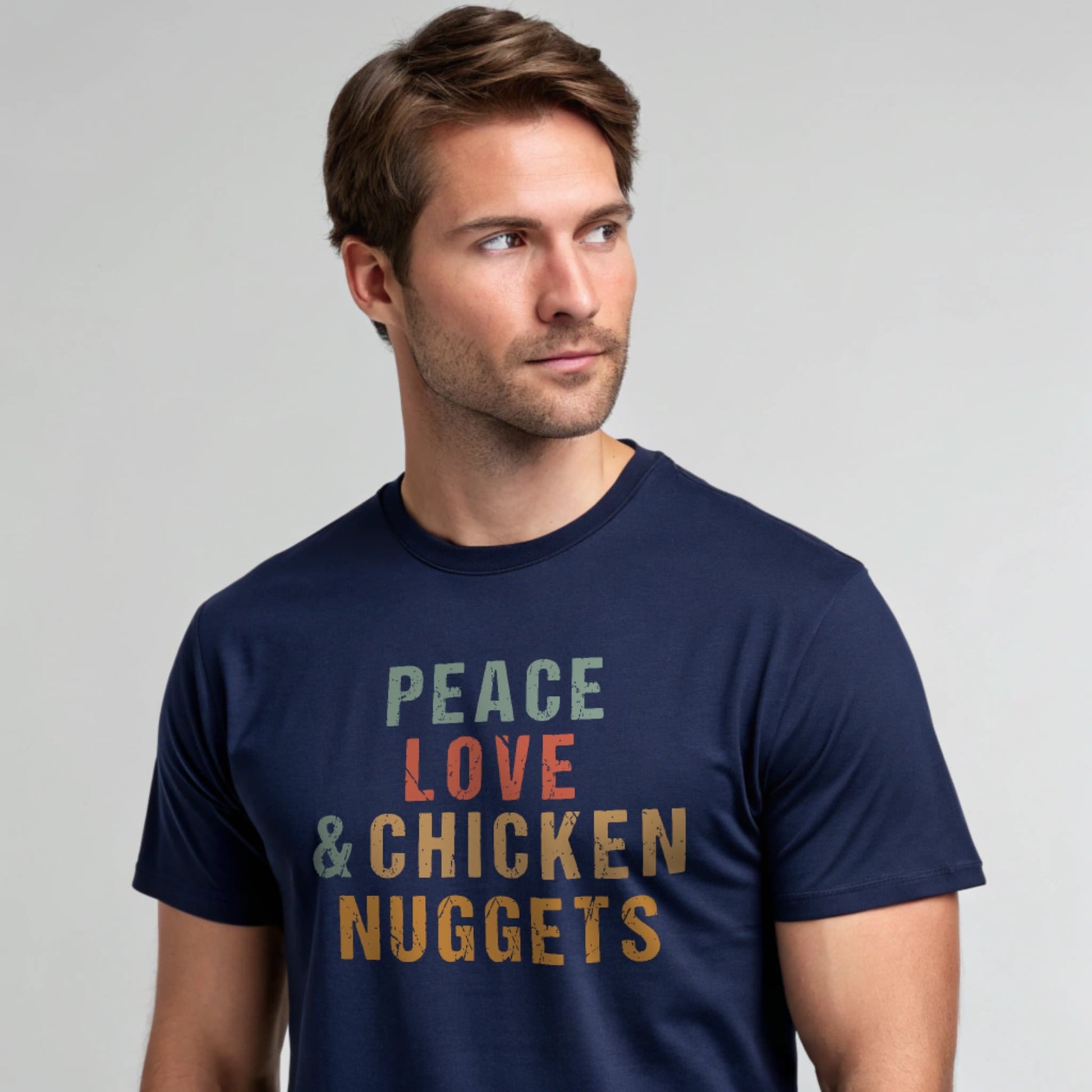 Male model wearing a navy crew-neck t-shirt with “Peace Love & Chicken Nuggets” text in multicolor block letters, studio portrait on a light-gray background.