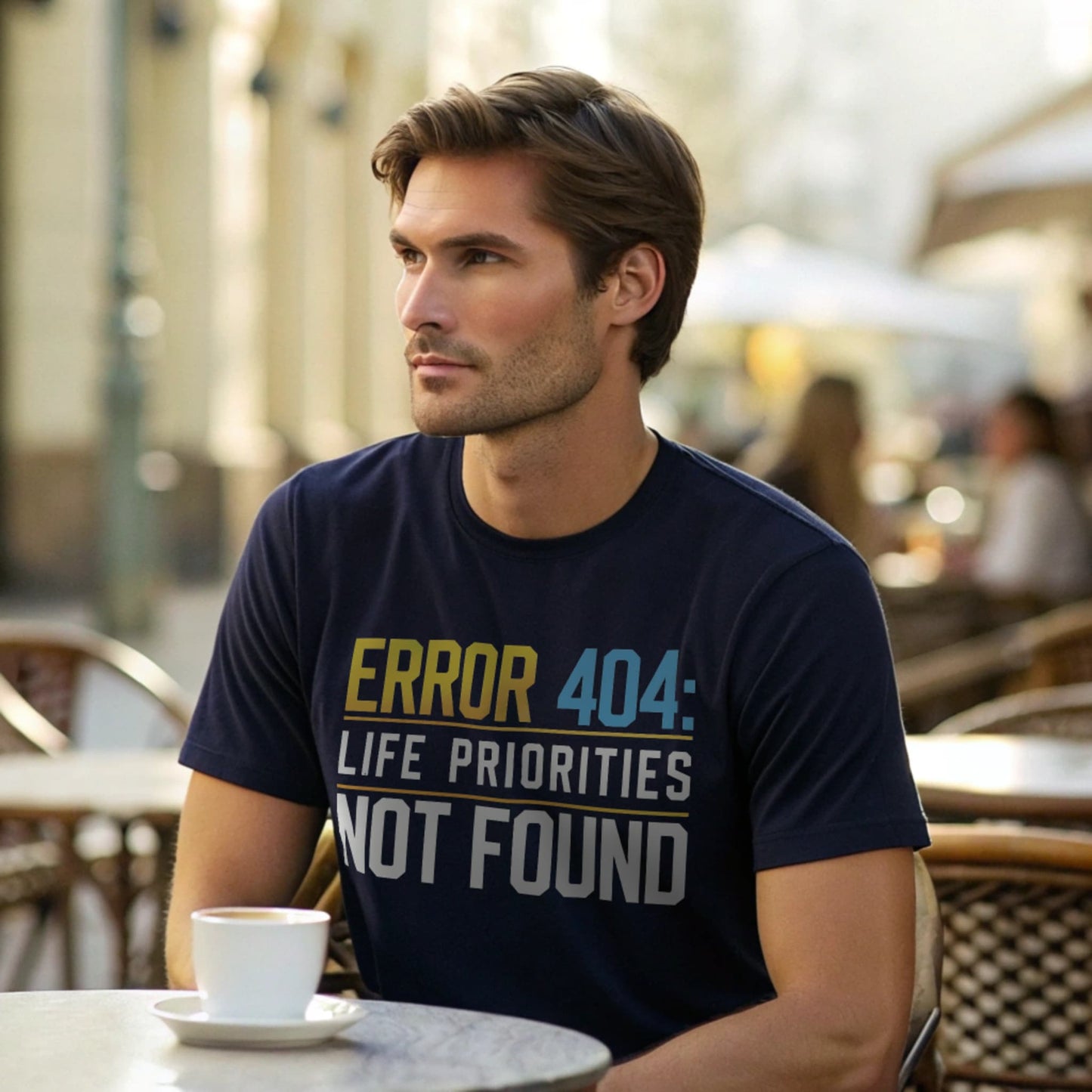 Male model seated at an outdoor café in a navy crew-neck t-shirt with “Priorities Not Found” tech graphic, lifestyle café shot.