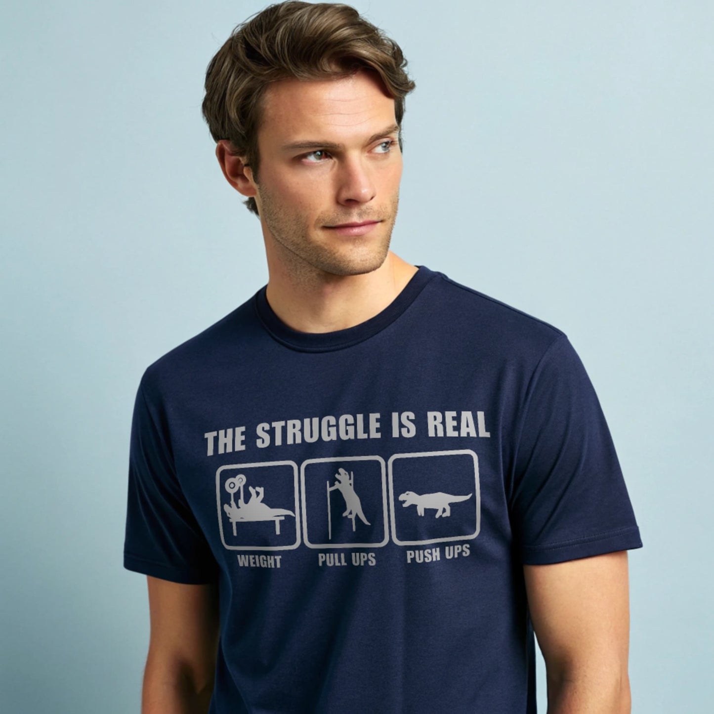 Male model wearing a navy crew-neck t-shirt with “The Struggle Is Real” graphic, studio shot on a pale blue background.