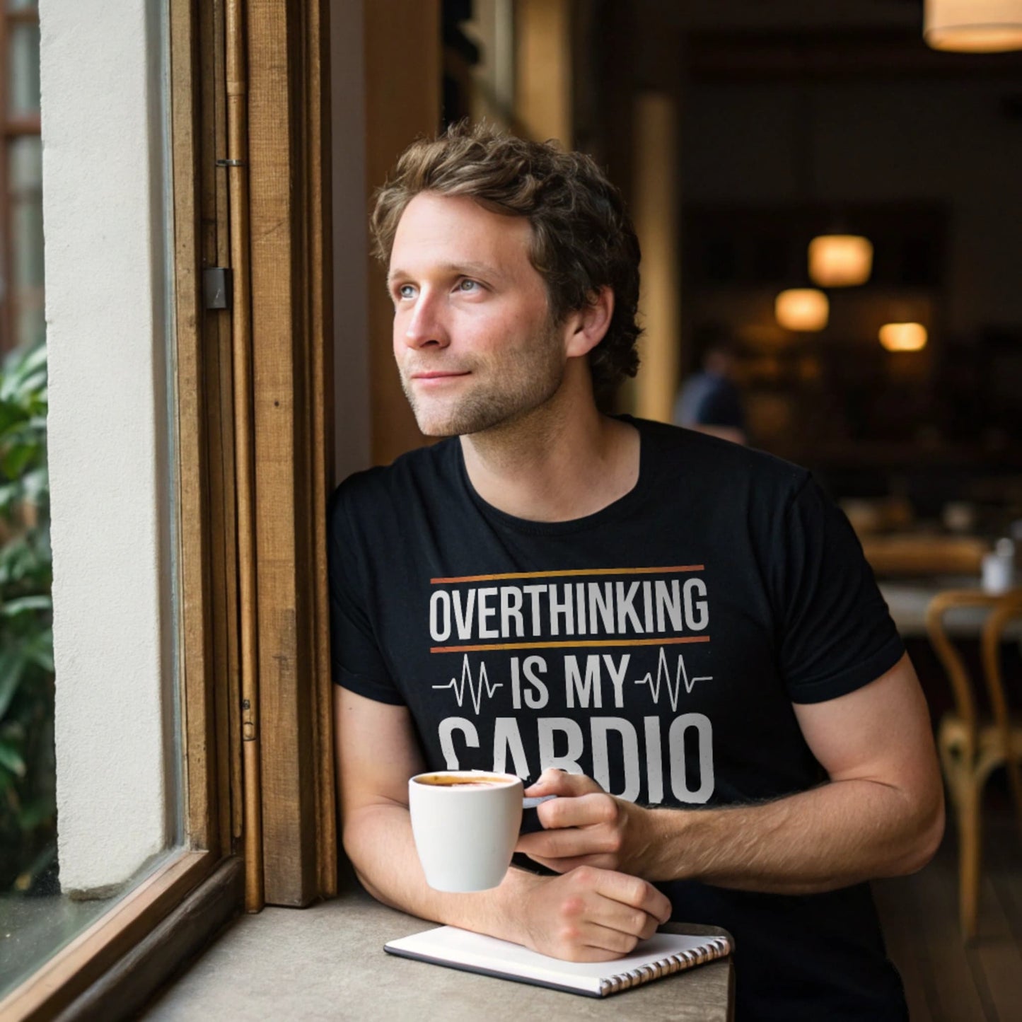 Hyper-realistic mockup of a 25–40-year-old man wearing a black Overthinking Cardio T-Shirt leaning against a café window sill while holding a coffee mug with a notebook beside him.