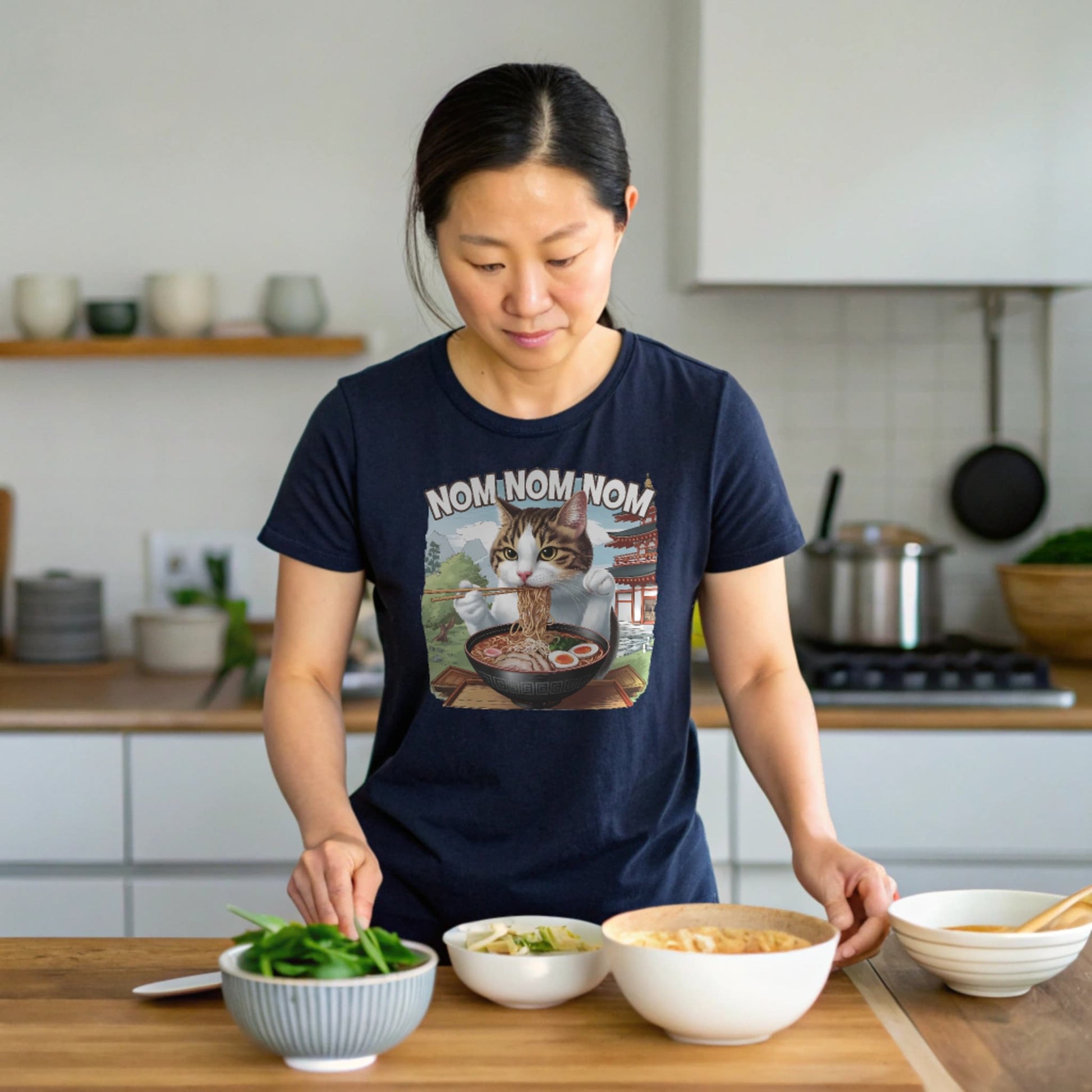 Navy lifestyle mockup of Ramen Cat T-Shirt on a female model at a home kitchen counter with ramen bowls, natural eyes