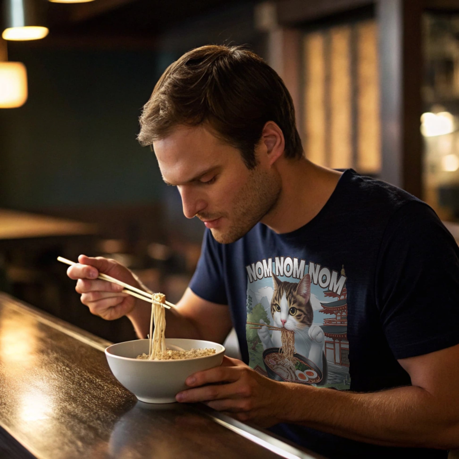 Navy lifestyle mockup of Ramen Cat T-Shirt on a male model slurping noodles at a ramen bar counter, warm ambient lighting