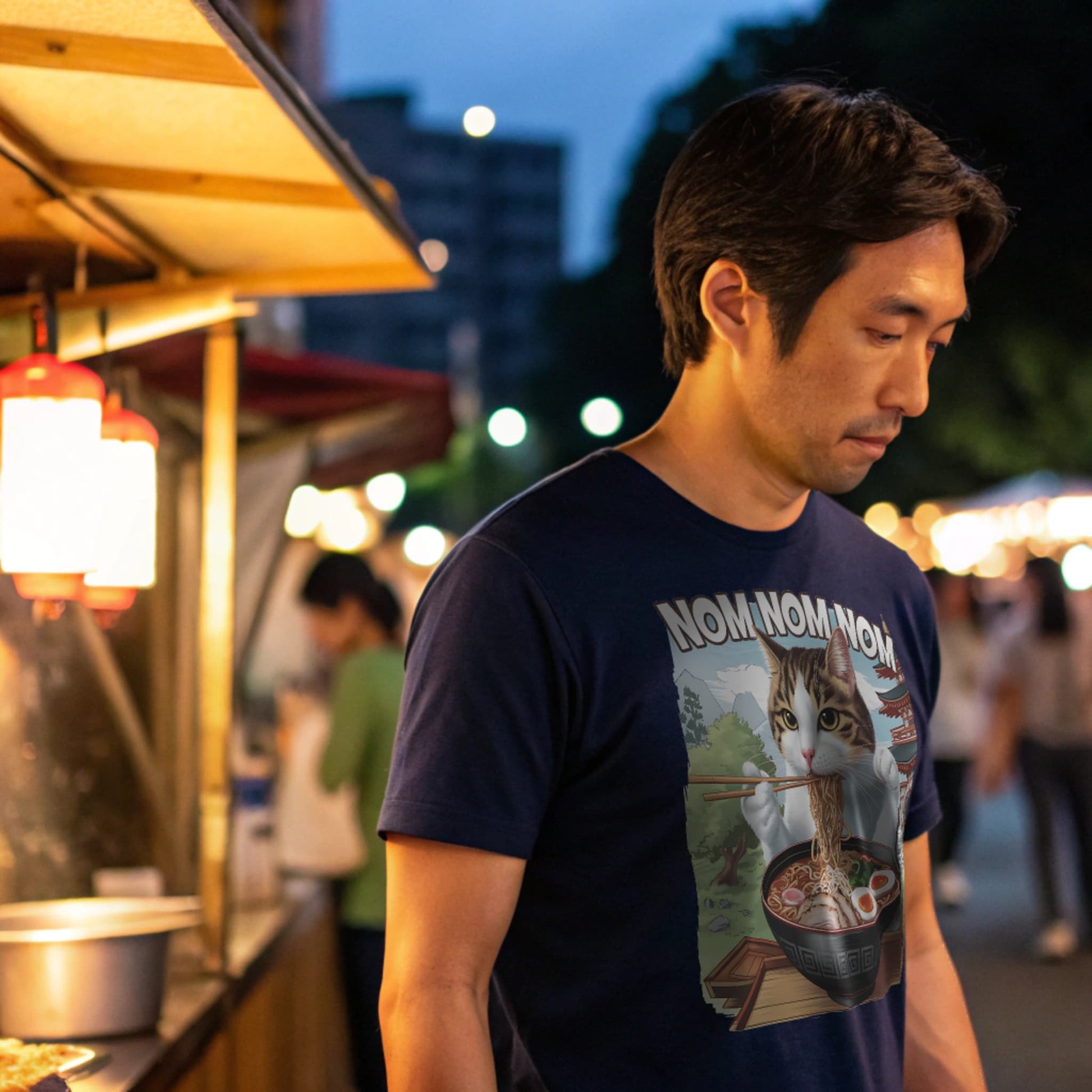 Navy lifestyle mockup of Ramen Cat T-Shirt on a male model ordering ramen at a street food stall under lantern lights