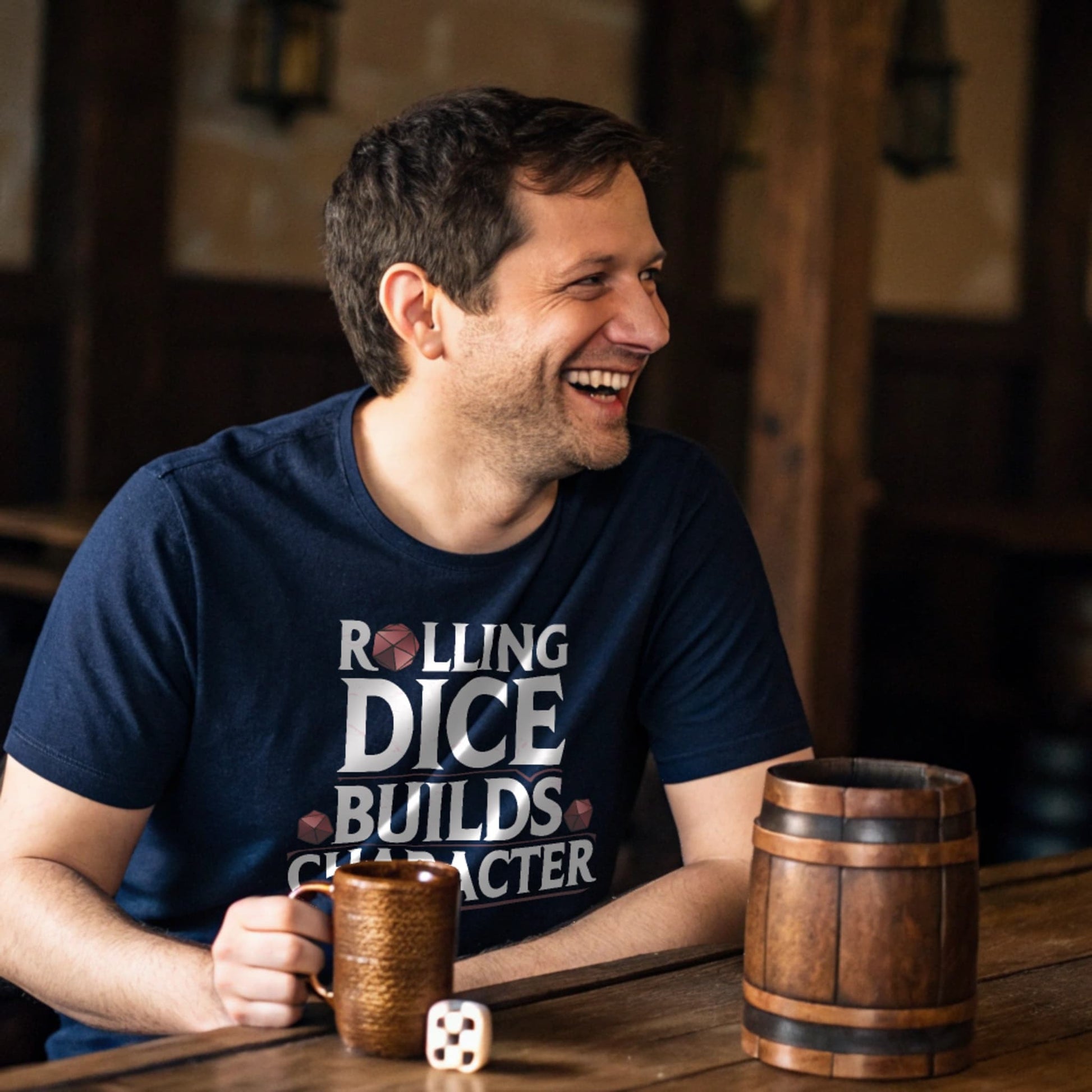 Navy lifestyle mockup of Rolling Dice Builds Character tee at a rustic tavern table with wooden mugs and dice, male model mid-laugh
