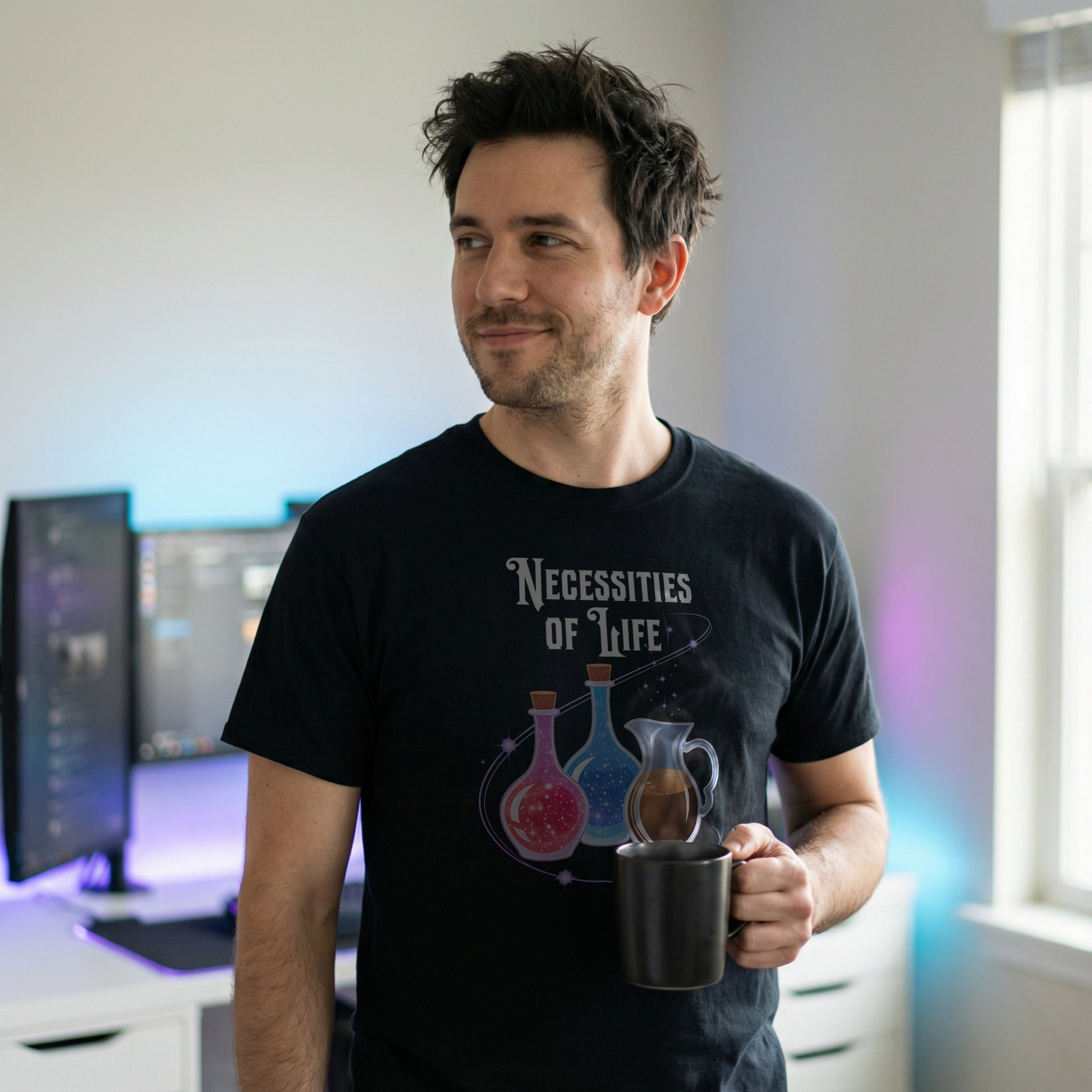 Man wearing a black Necessities of Life T-shirt holding a coffee mug in a workstation setup