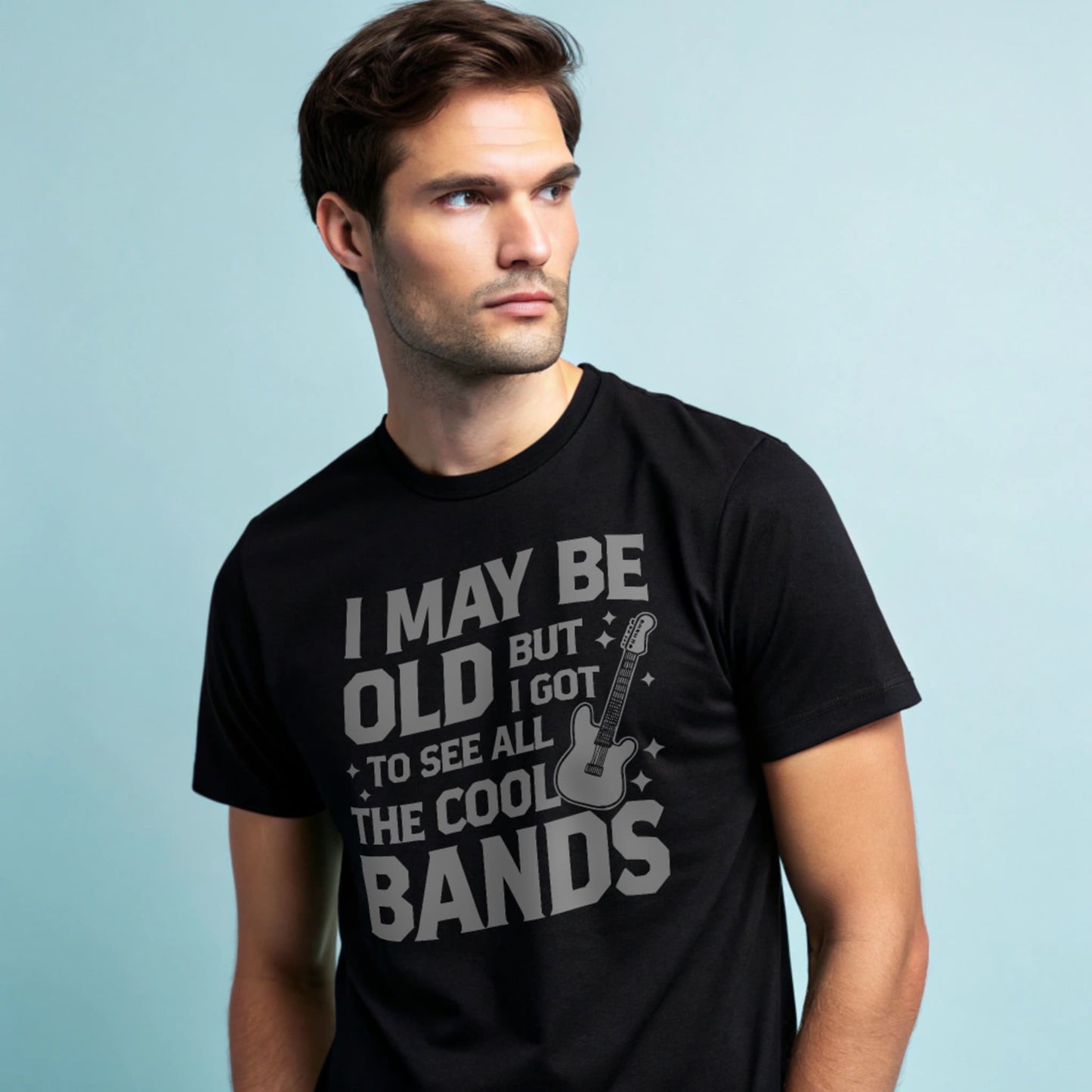 Male model wearing a black crew-neck t-shirt with a vintage guitar graphic and “I May Be Old But I Got To See All The Cool Bands” text, studio shot on a smooth pale-blue background.