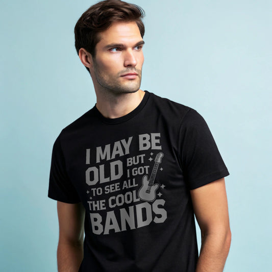 Male model wearing a black crew-neck t-shirt with a vintage guitar graphic and “I May Be Old But I Got To See All The Cool Bands” text, studio shot on a smooth pale-blue background.