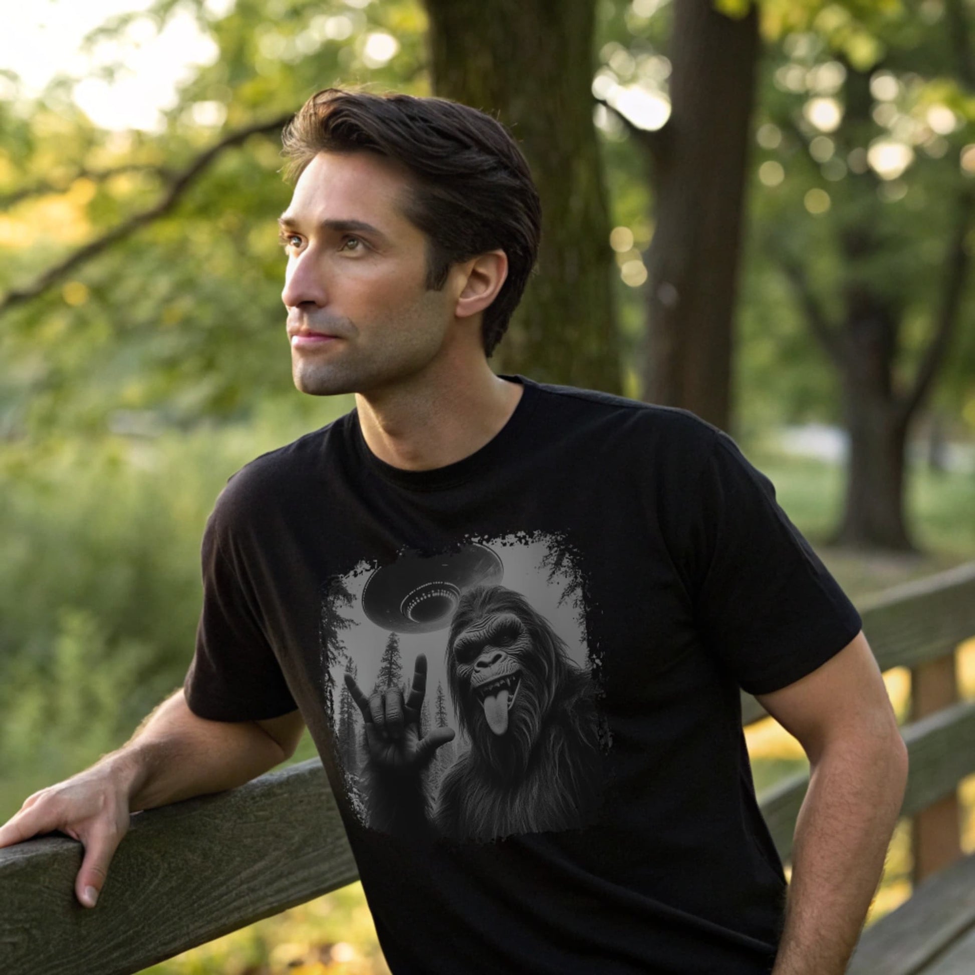 Male model leaning against a wooden bench wearing a black crew-neck t-shirt featuring a white silhouette of Sasquatch raising its hand graphic, lifestyle park shot.