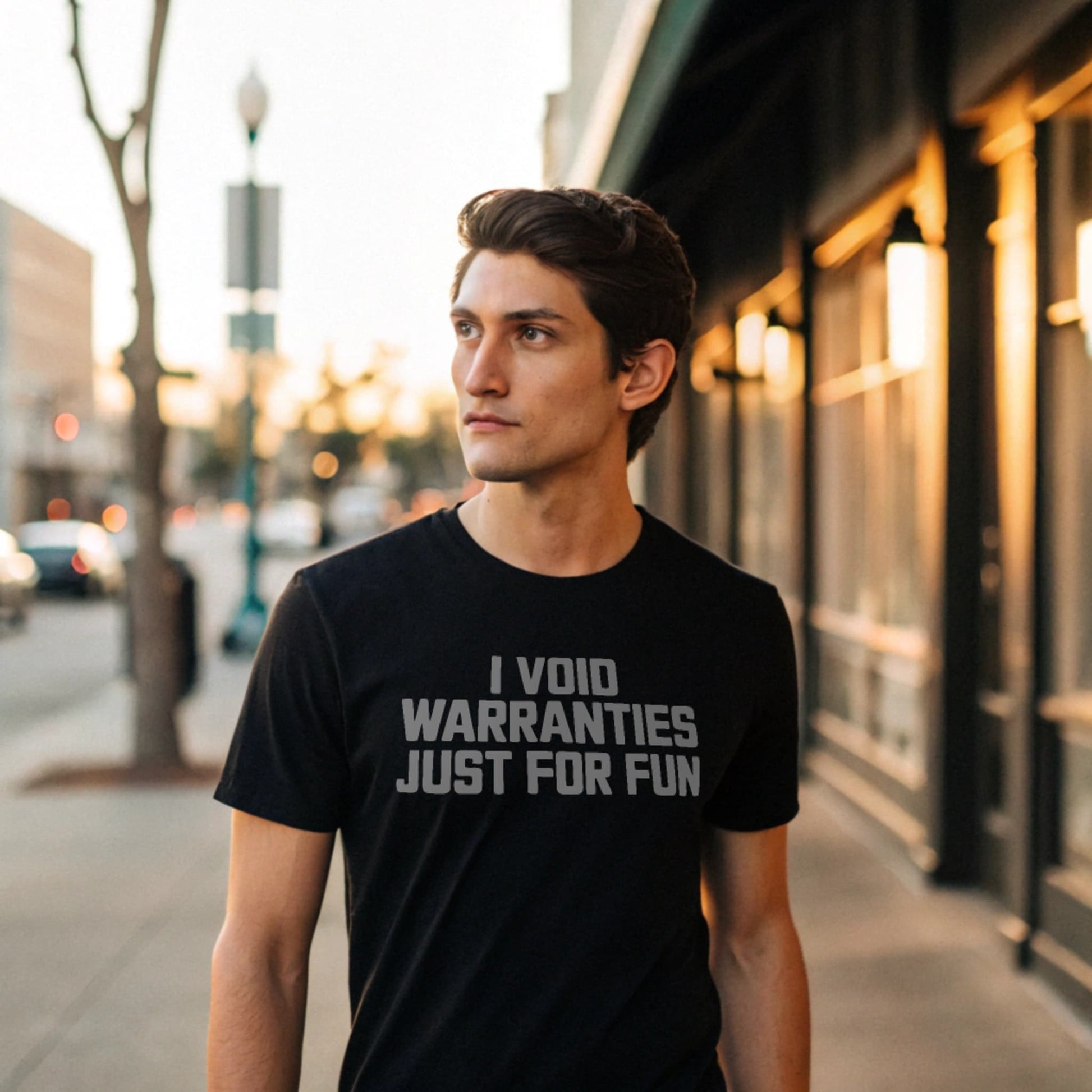 Male model strolling an urban sidewalk in a plain black Gildan 64000 crew-neck t-shirt with “I VOID WARRANTIES JUST FOR FUN” text, street-style lifestyle shot.