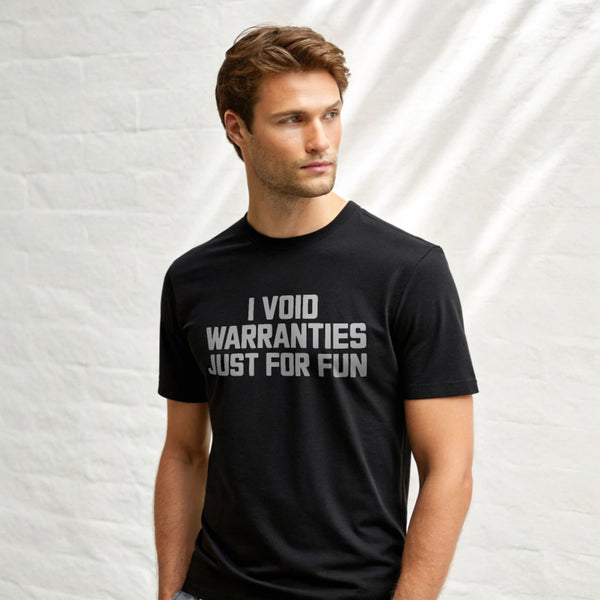 Male model wearing a plain black Gildan 64000 crew-neck t-shirt with “I VOID WARRANTIES JUST FOR FUN” text, studio shot on white background.