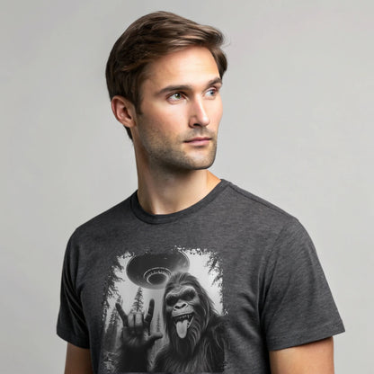 Male model wearing a charcoal crew-neck t-shirt featuring a white silhouette of Sasquatch raising its hand graphic, studio shot on a neutral gray backdrop.