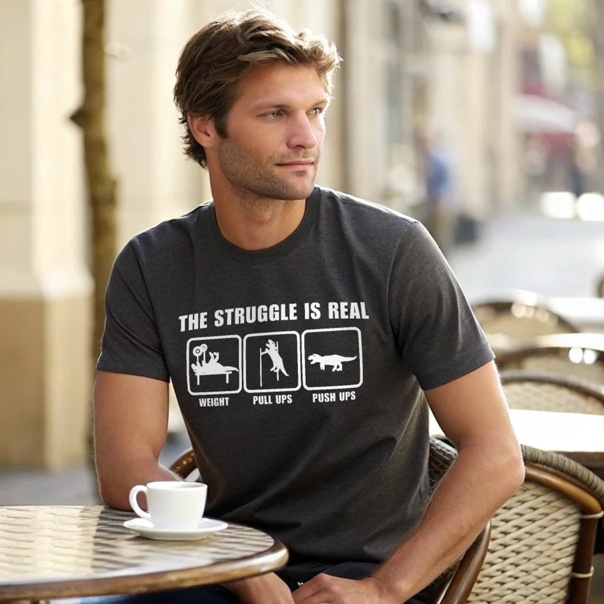 Male model seated at an outdoor café table in a charcoal crew-neck t-shirt with “The Struggle Is Real” graphic, lifestyle café shot.