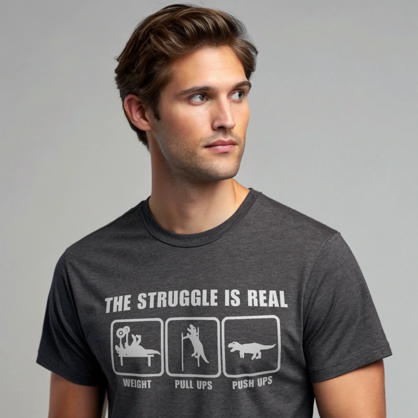 Male model wearing a charcoal crew-neck t-shirt with “The Struggle Is Real” graphic, studio portrait against a gray background.