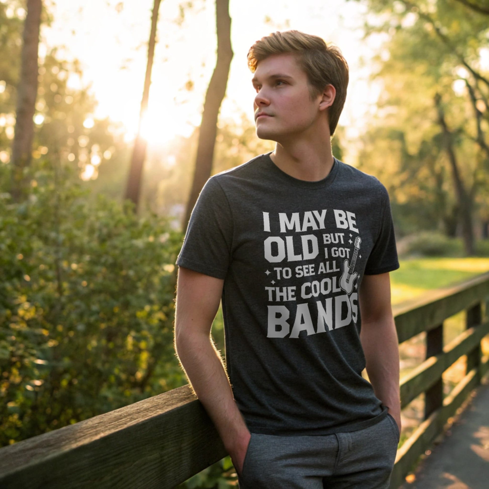 Male model walking through a sunlit park wearing a dark heather crew-neck t-shirt with a vintage guitar graphic and “I May Be Old But I Got To See All The Cool Bands” text, lifestyle outdoor shot.