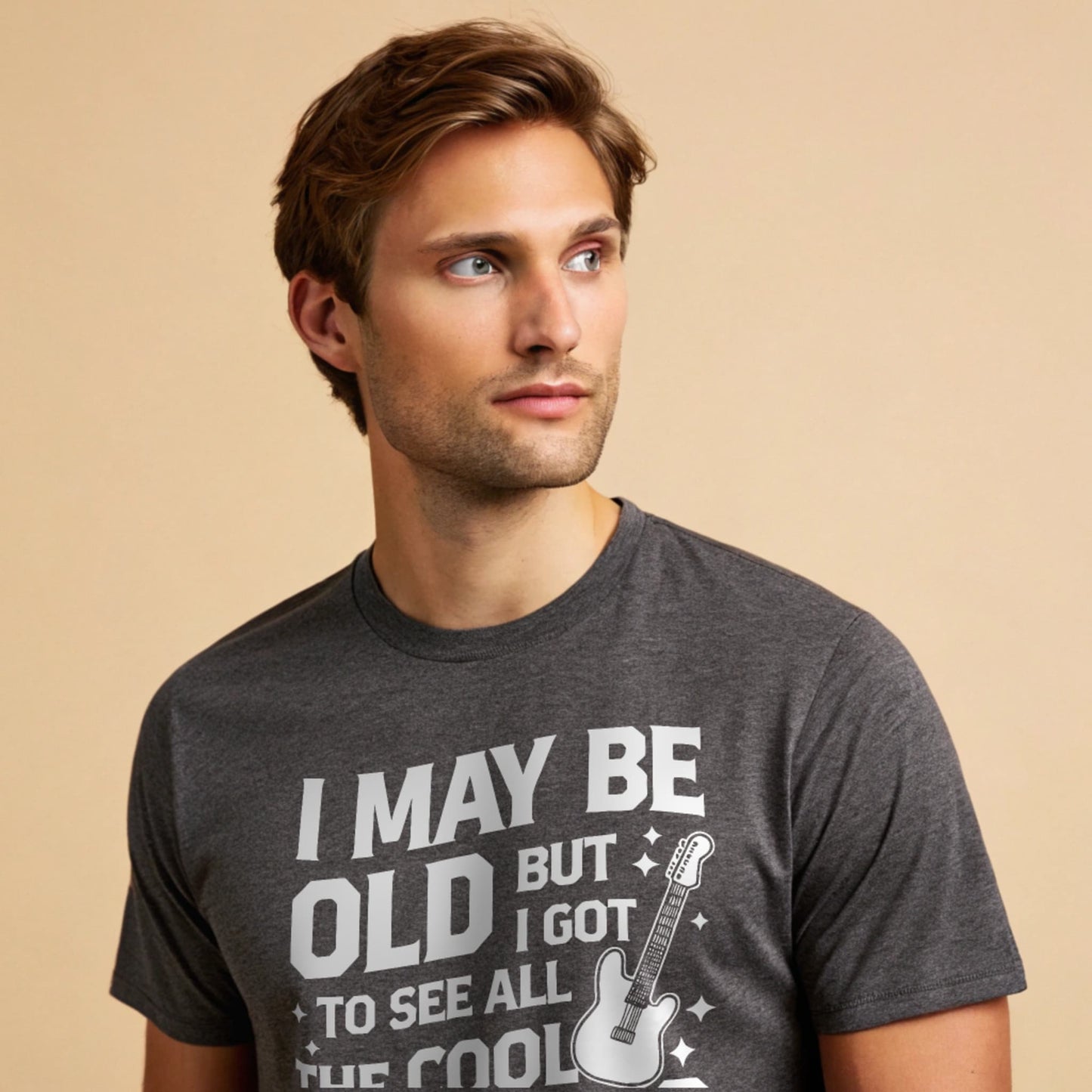 Male model wearing a dark heather crew-neck t-shirt with a vintage guitar graphic and “I May Be Old But I Got To See All The Cool Bands” text, studio shot on a smooth warm-beige background.