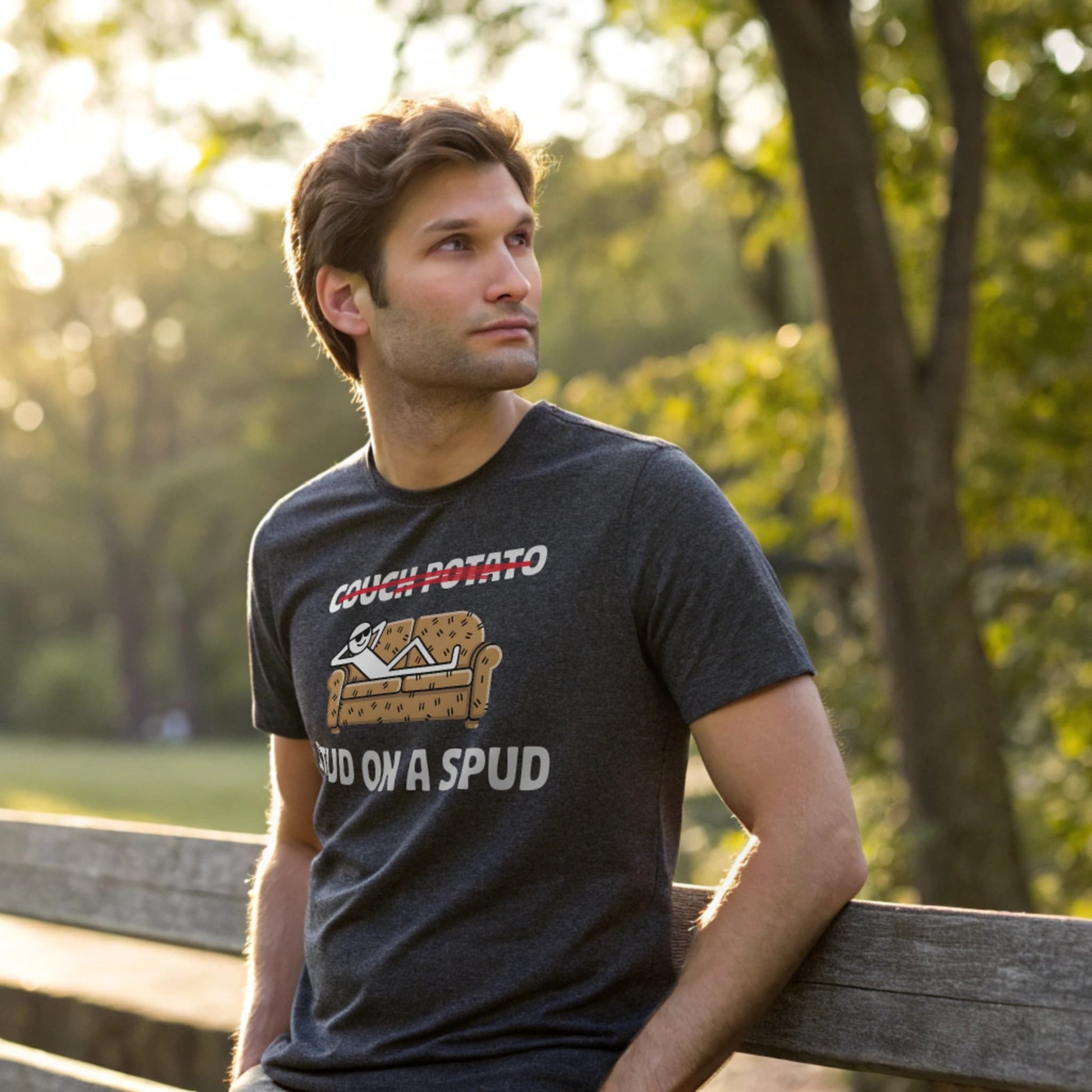 Male model leaning on a wooden bench in a dark heather crew-neck t-shirt with “Couch Potato: Stud on a Spud” graphic, lifestyle park shot.