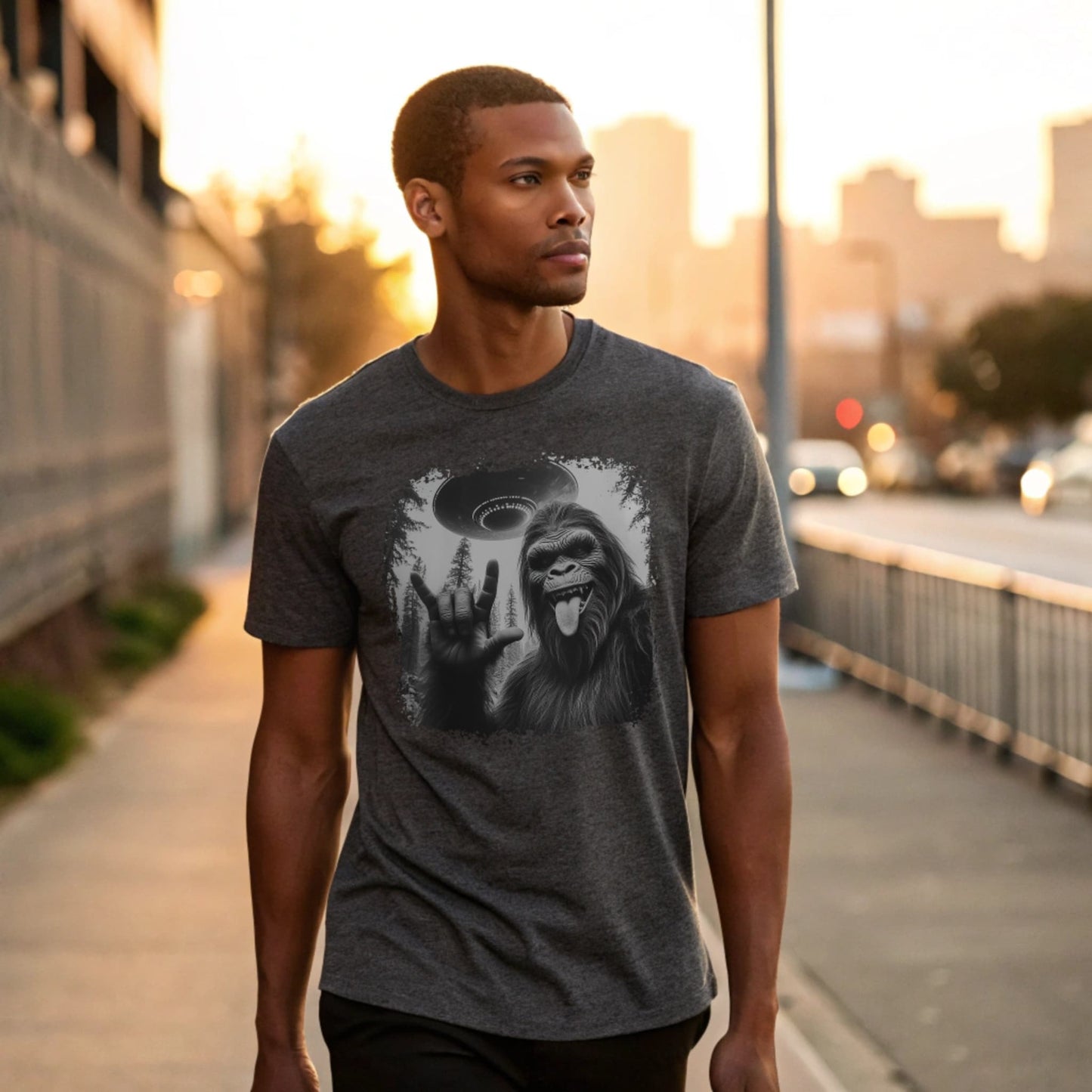 Male model strolling an urban sidewalk wearing a dark heather crew-neck t-shirt featuring a white silhouette of Sasquatch raising its hand graphic, lifestyle street shot.