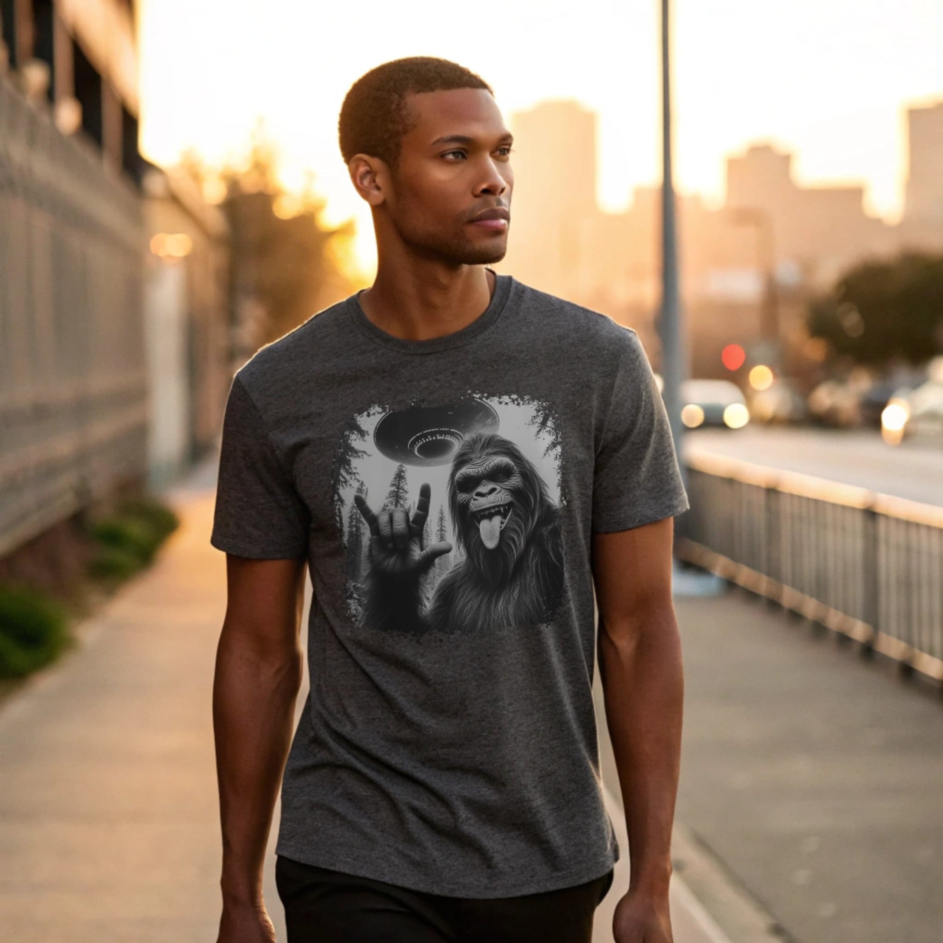 Male model strolling an urban sidewalk wearing a dark heather crew-neck t-shirt featuring a white silhouette of Sasquatch raising its hand graphic, lifestyle street shot.