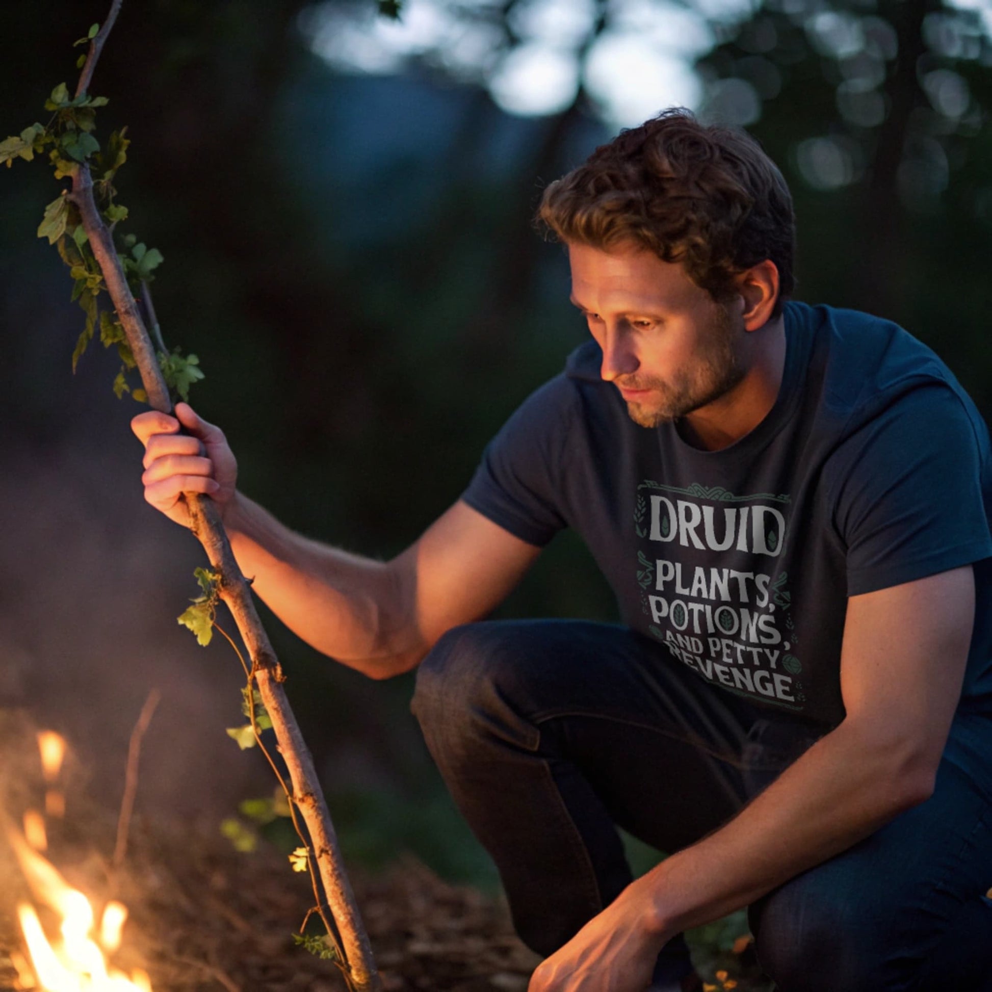 Navy lifestyle mockup of Druid Petty Revenge T-Shirt on a male model crouched by a glowing campfire stirring a leafy staff, plain crew neck