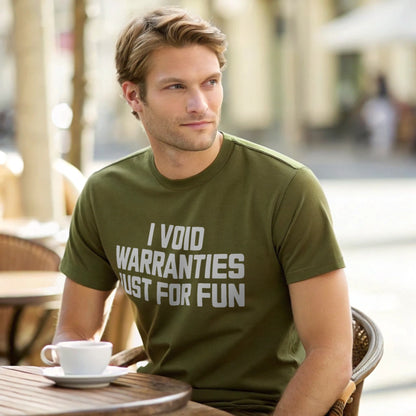 Male model seated at an outdoor café table wearing a plain military green Gildan 64000 crew-neck t-shirt with “I VOID WARRANTIES JUST FOR FUN” text, natural-light lifestyle shot.