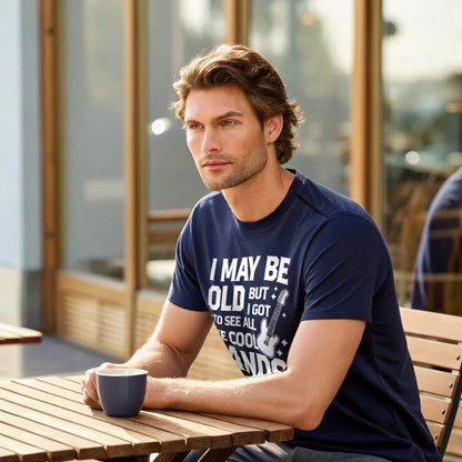 Male model seated at an outdoor café wearing a navy crew-neck t-shirt with a vintage guitar graphic and “I May Be Old But I Got To See All The Cool Bands” text, lifestyle café shot.
