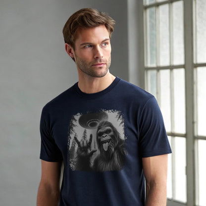 Male model wearing a navy crew-neck t-shirt featuring a white silhouette of Sasquatch raising its hand graphic, studio shot by a window-lit backdrop.
