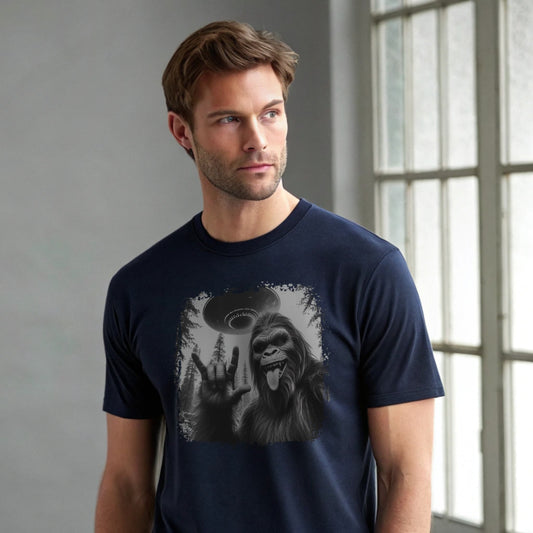 Male model wearing a navy crew-neck t-shirt featuring a white silhouette of Sasquatch raising its hand graphic, studio shot by a window-lit backdrop.