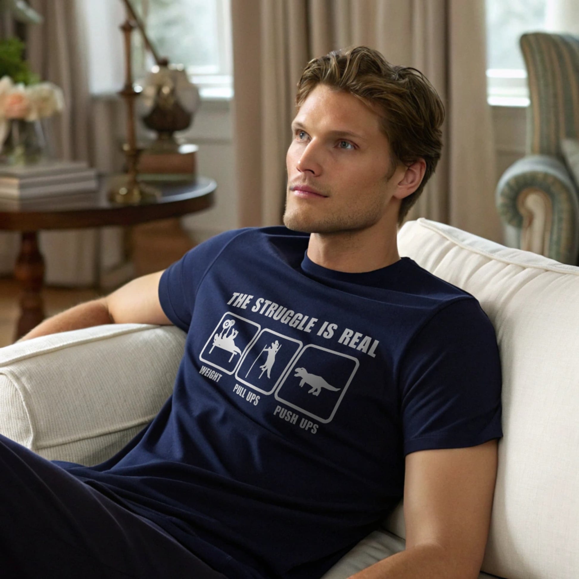 Male model lounging in a living room setting wearing a navy crew-neck t-shirt with “The Struggle Is Real” graphic, lifestyle home interior shot.