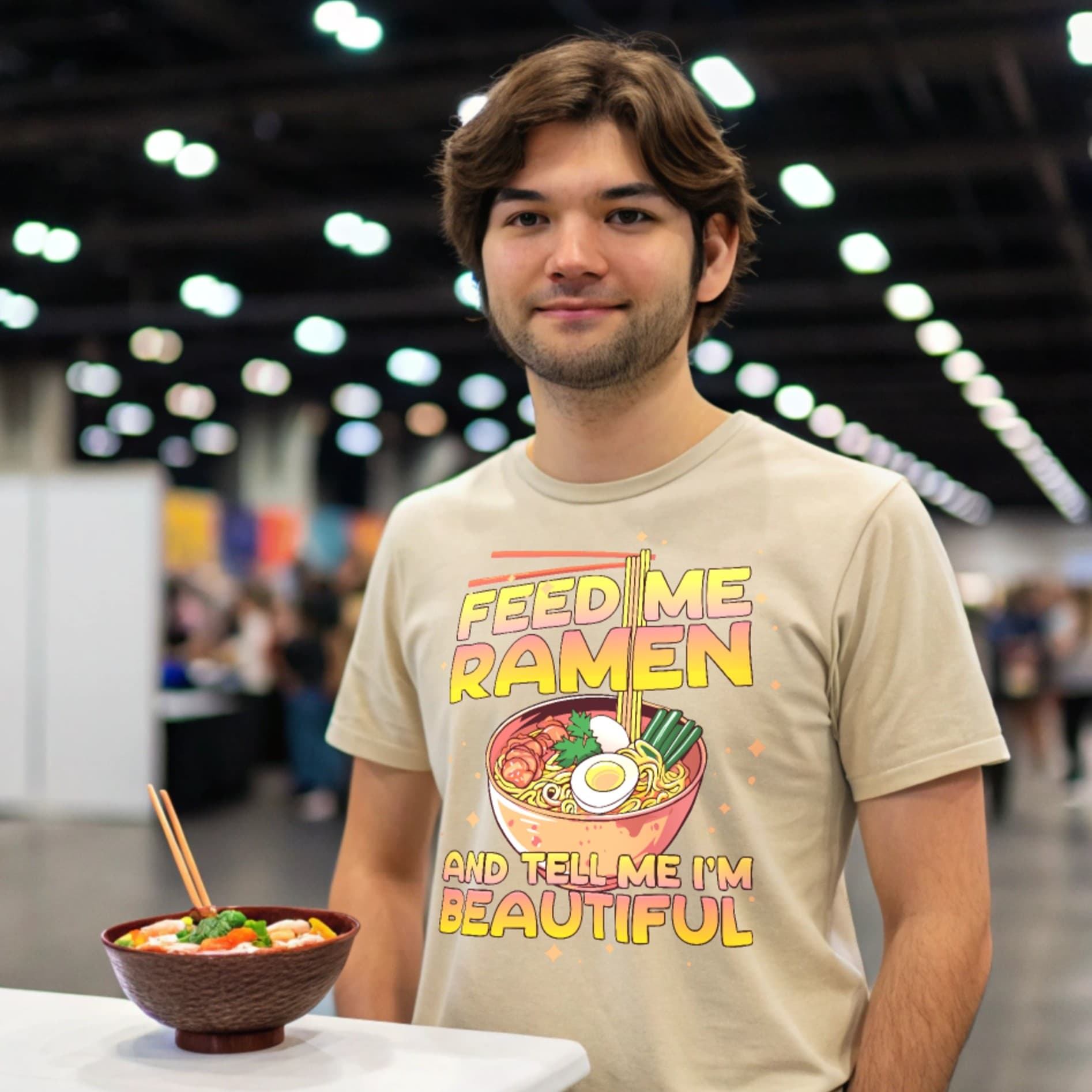 Otaku at a convention wearing a natural-colored Ramen Lover T-Shirt beside a ramen bowl.