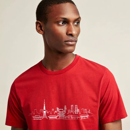 Male model wearing a red crew-neck t-shirt with a white city skyline graphic, studio portrait on a pale-cream background.